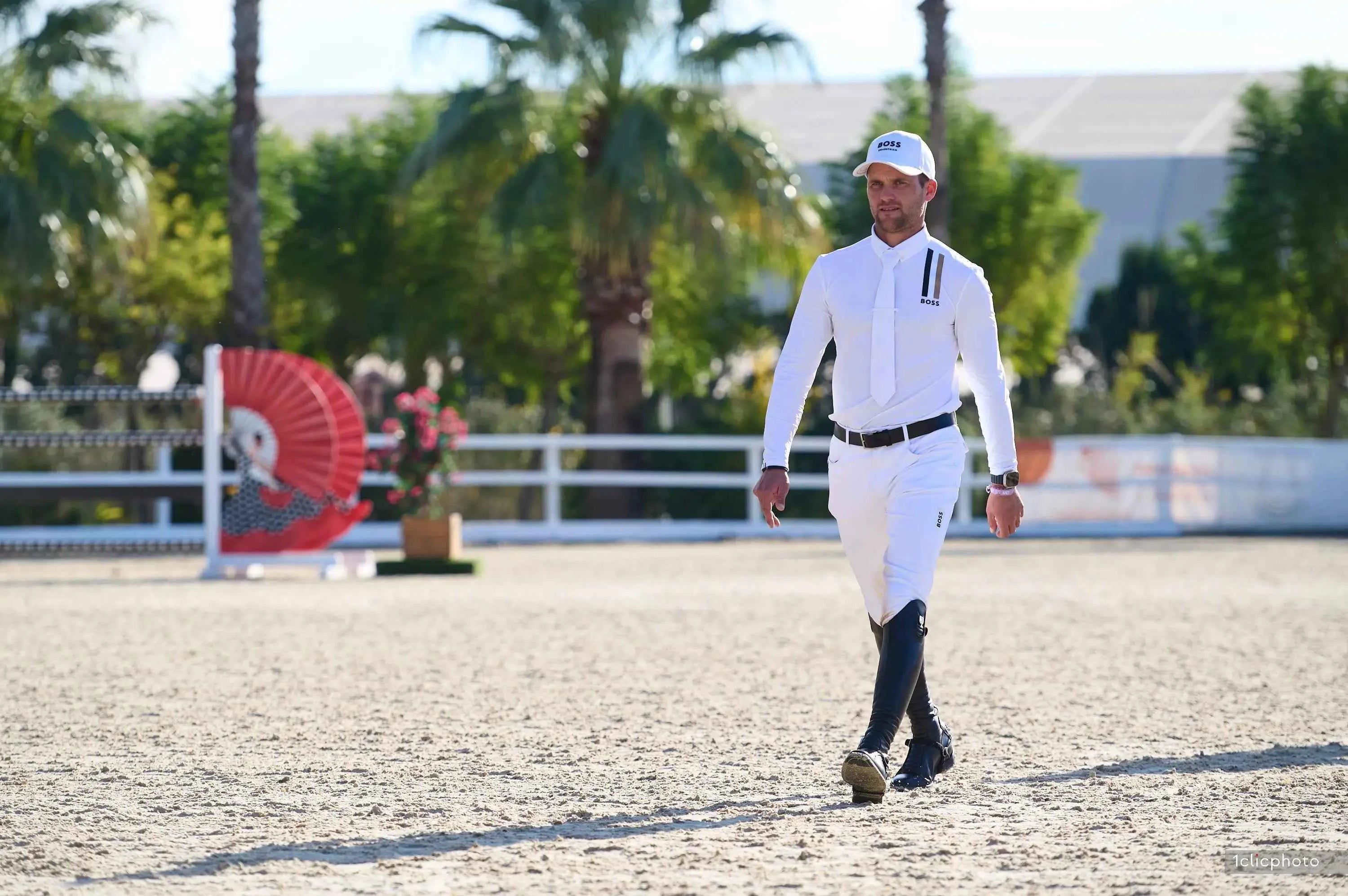Alexander Schill in equestrian attire walking on a dirt path with palm trees in the background