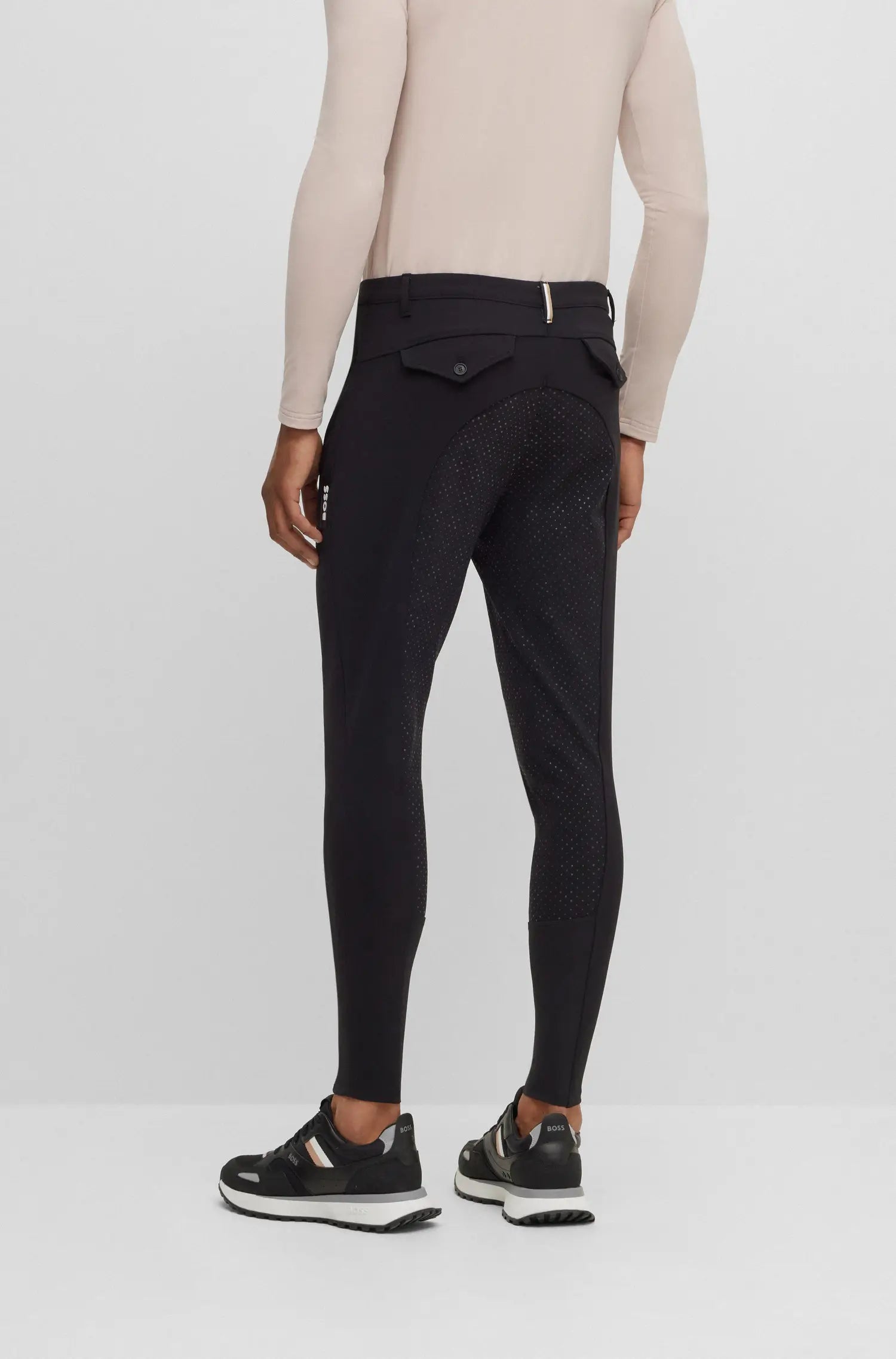 Black | A model from the back wearing black equestrian riding tights with a textured pattern, paired with a beige long-sleeve top and black sneakers, against a light gray studio background.