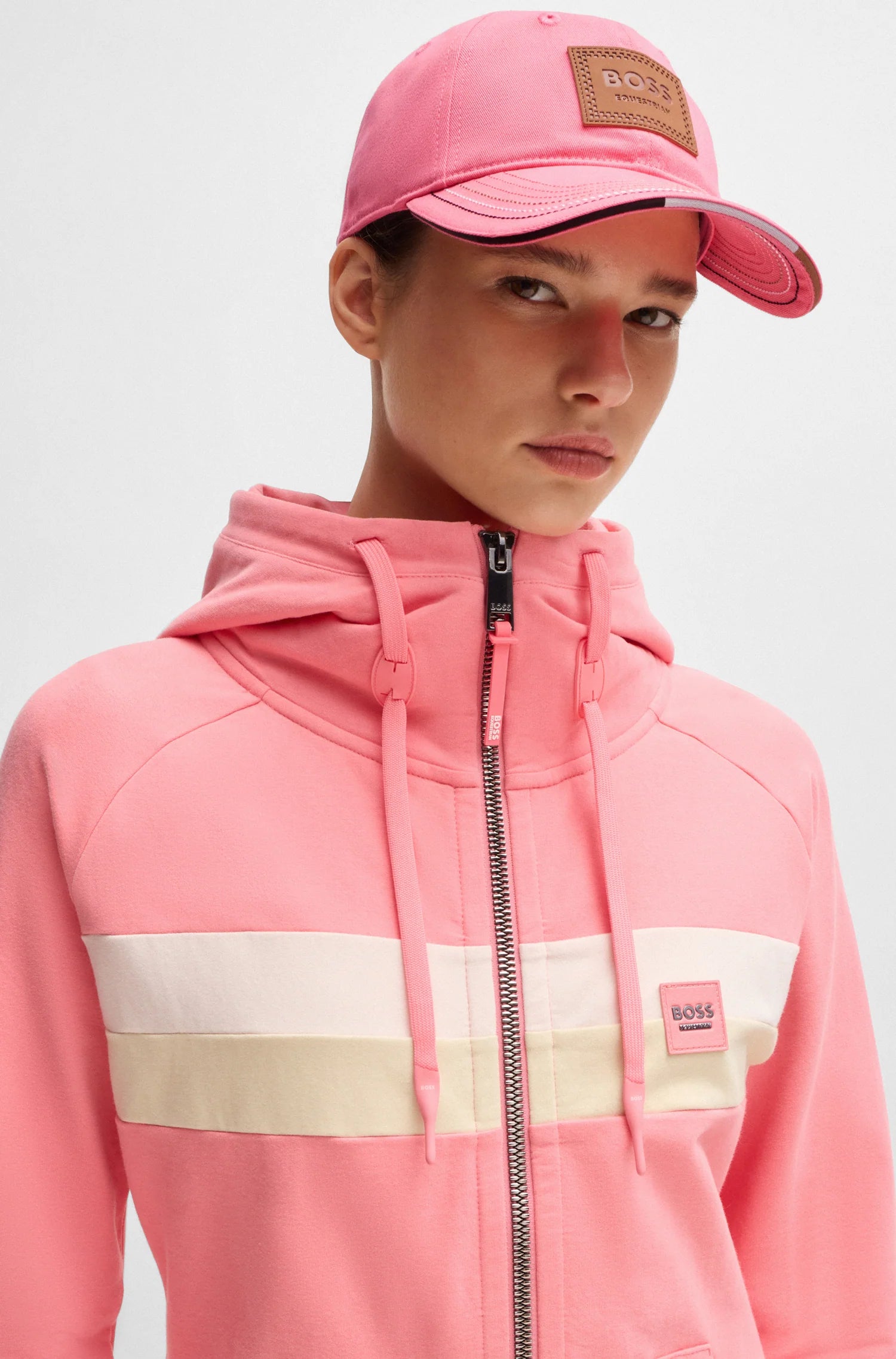 Pink | A model wearing a pink zip-up hoodie with cream color-blocked stripes across the chest, a matching pink BOSS cap, and a pink BOSS wrist strap, photographed against a light gray studio background.