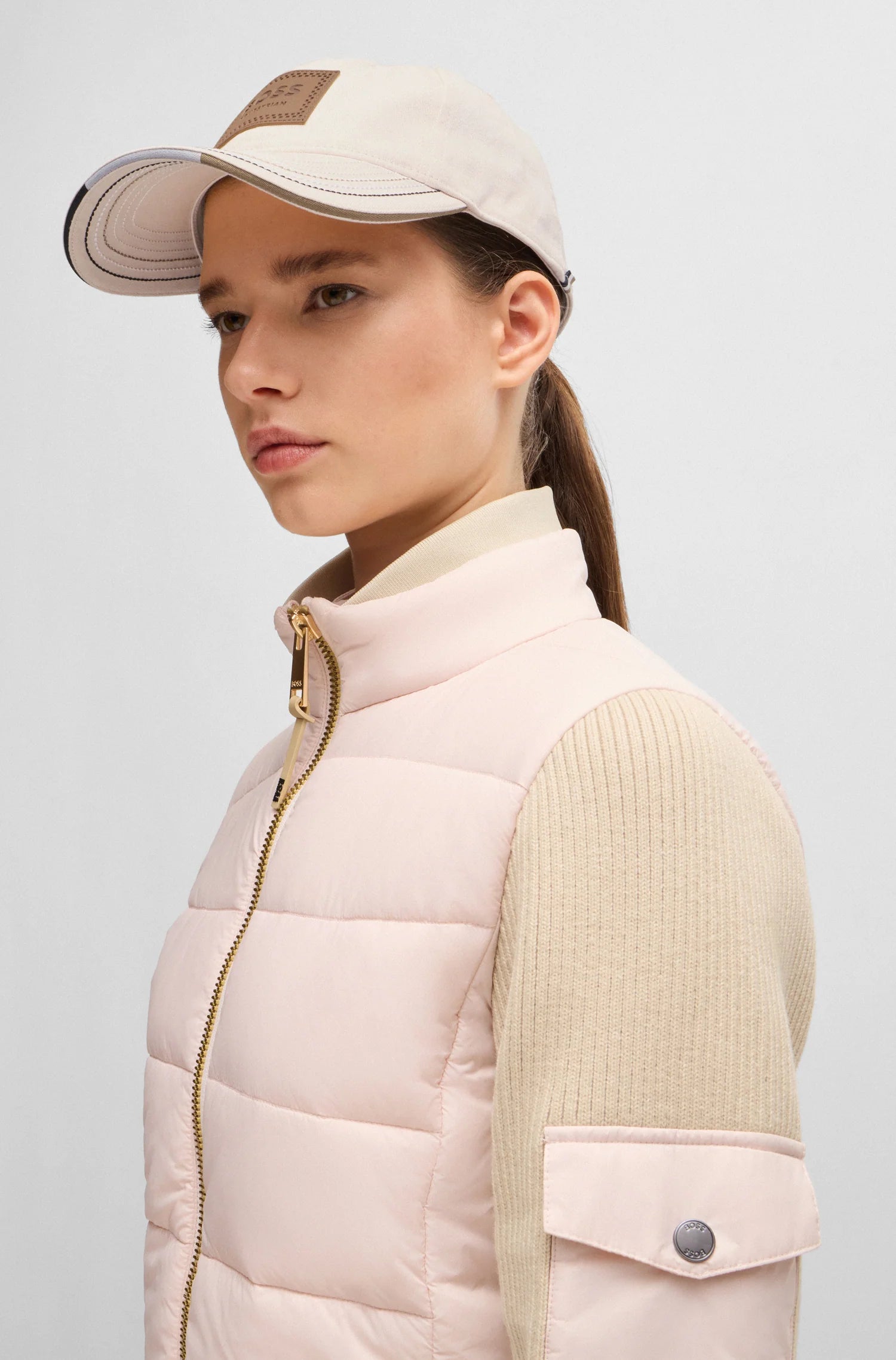 Pearl | A woman in profile wearing a pale pink puffer vest with beige ribbed knit sleeves, a cream baseball cap, and a cream mock neck top, photographed against a light gray studio background.