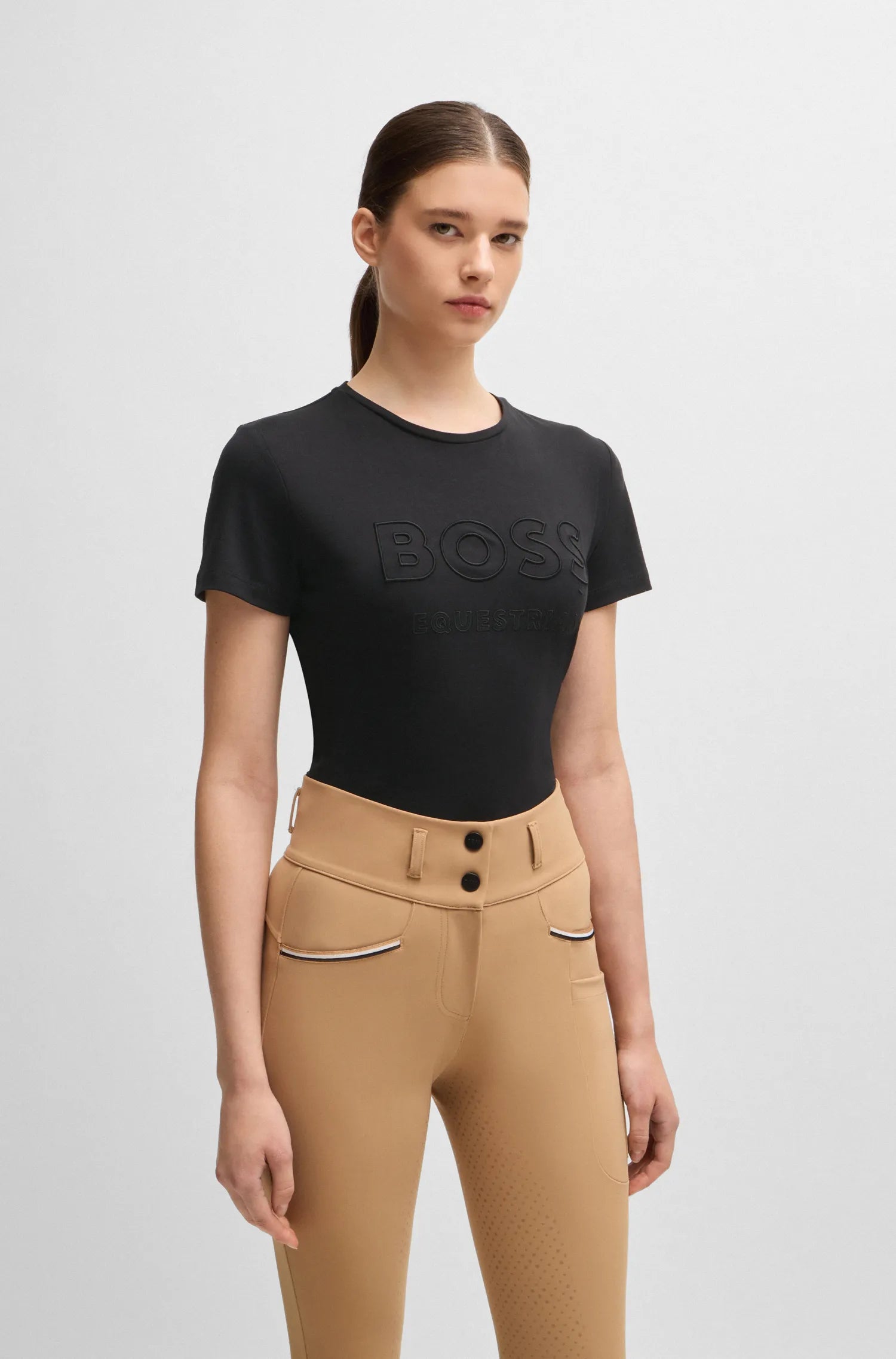 Black | A woman modeling a black short-sleeve BOSS T-shirt with tonal embossed lettering, paired with tan riding breeches, against a light gray studio background.