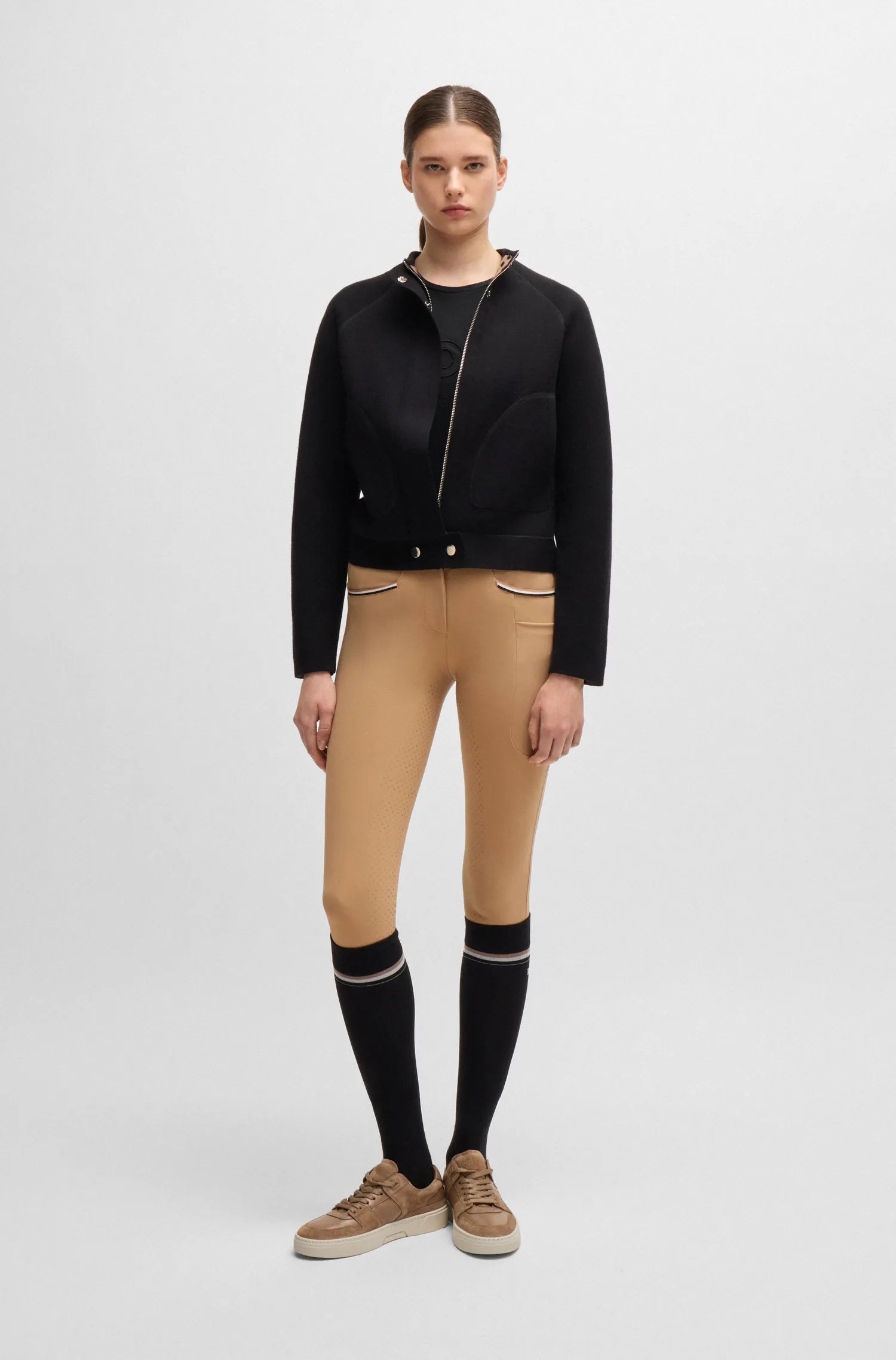 Black | A model stands against a light gray studio background wearing a black zip-front jacket, tan fitted pants, black knee-high socks with white stripes, and tan sneakers.