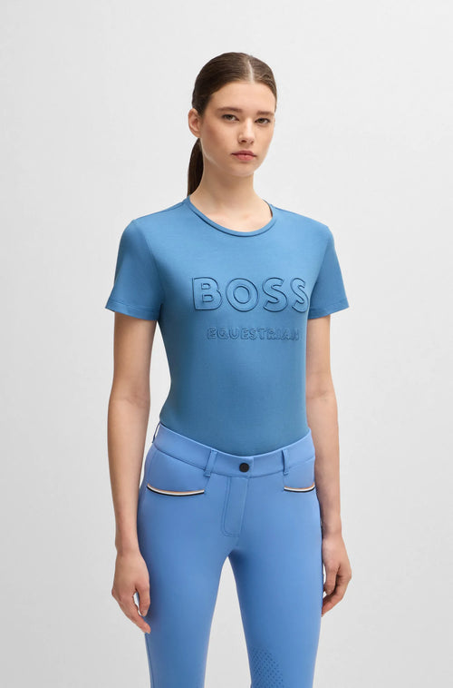 Riviera | A woman modeling a light blue short-sleeve t-shirt with 'BOSS' and 'HUGO BOSS' embossed on the chest, paired with matching blue riding breeches, against a light gray studio background.