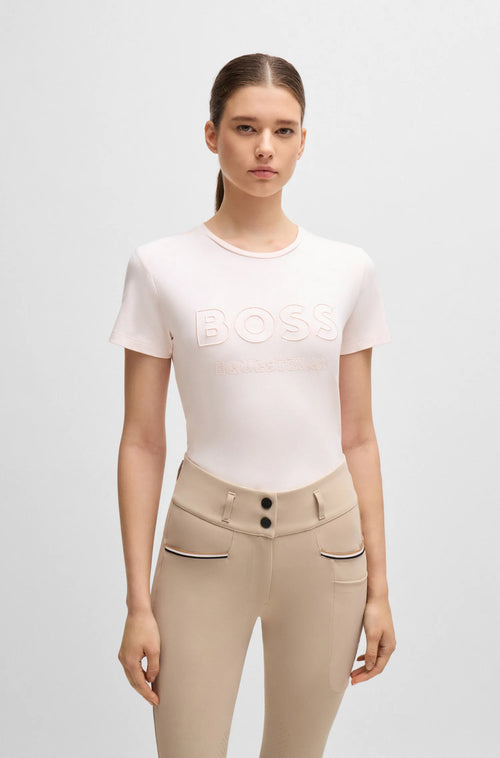 Pearl | A model wearing a light pink short-sleeve BOSS t-shirt and beige riding breeches with black buttons and striped pocket trim, standing against a light gray studio background.