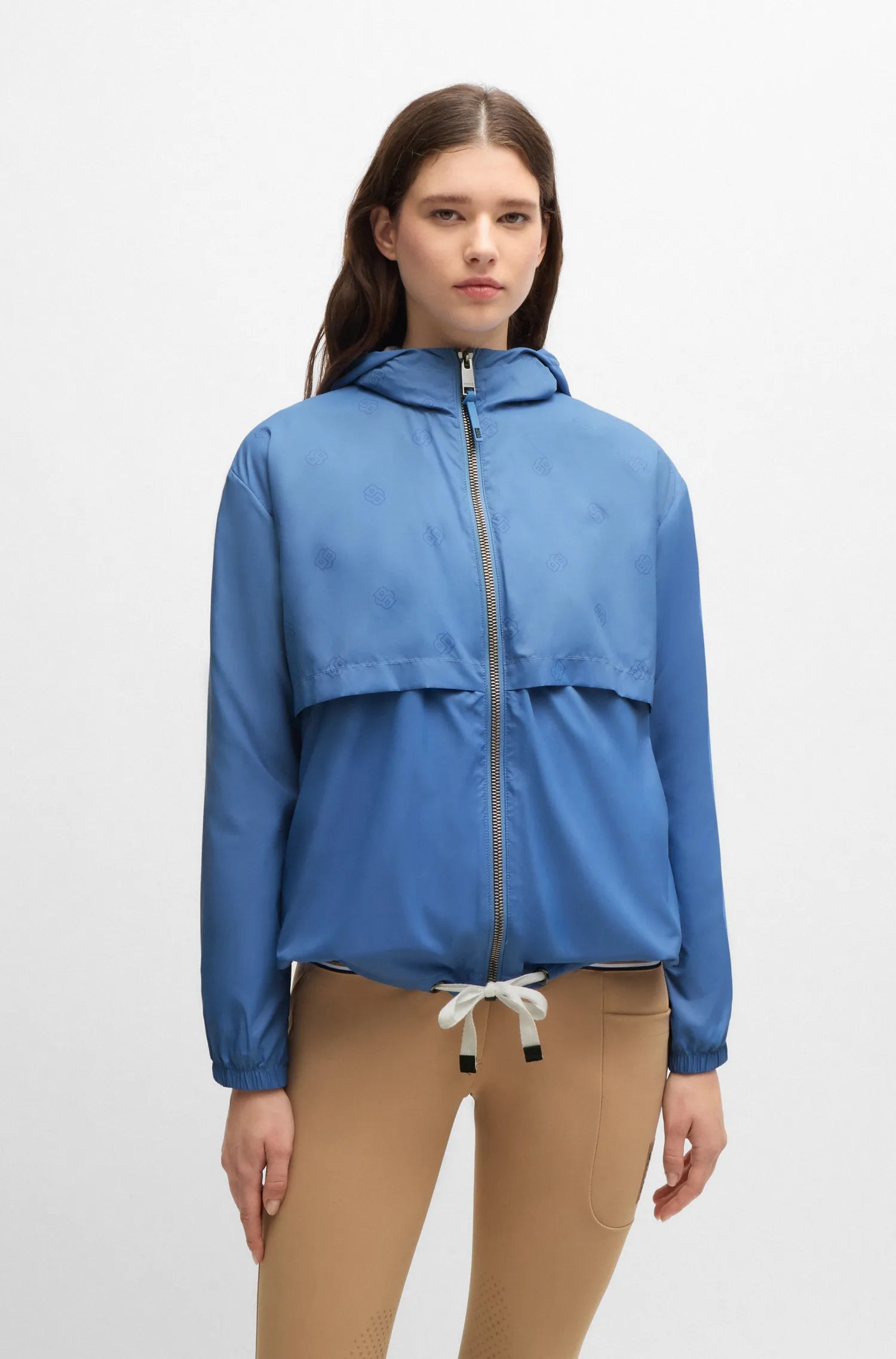 Riviera | A woman modeling a blue lightweight hooded jacket with a front zipper, paired with tan pants, against a plain light gray studio background.