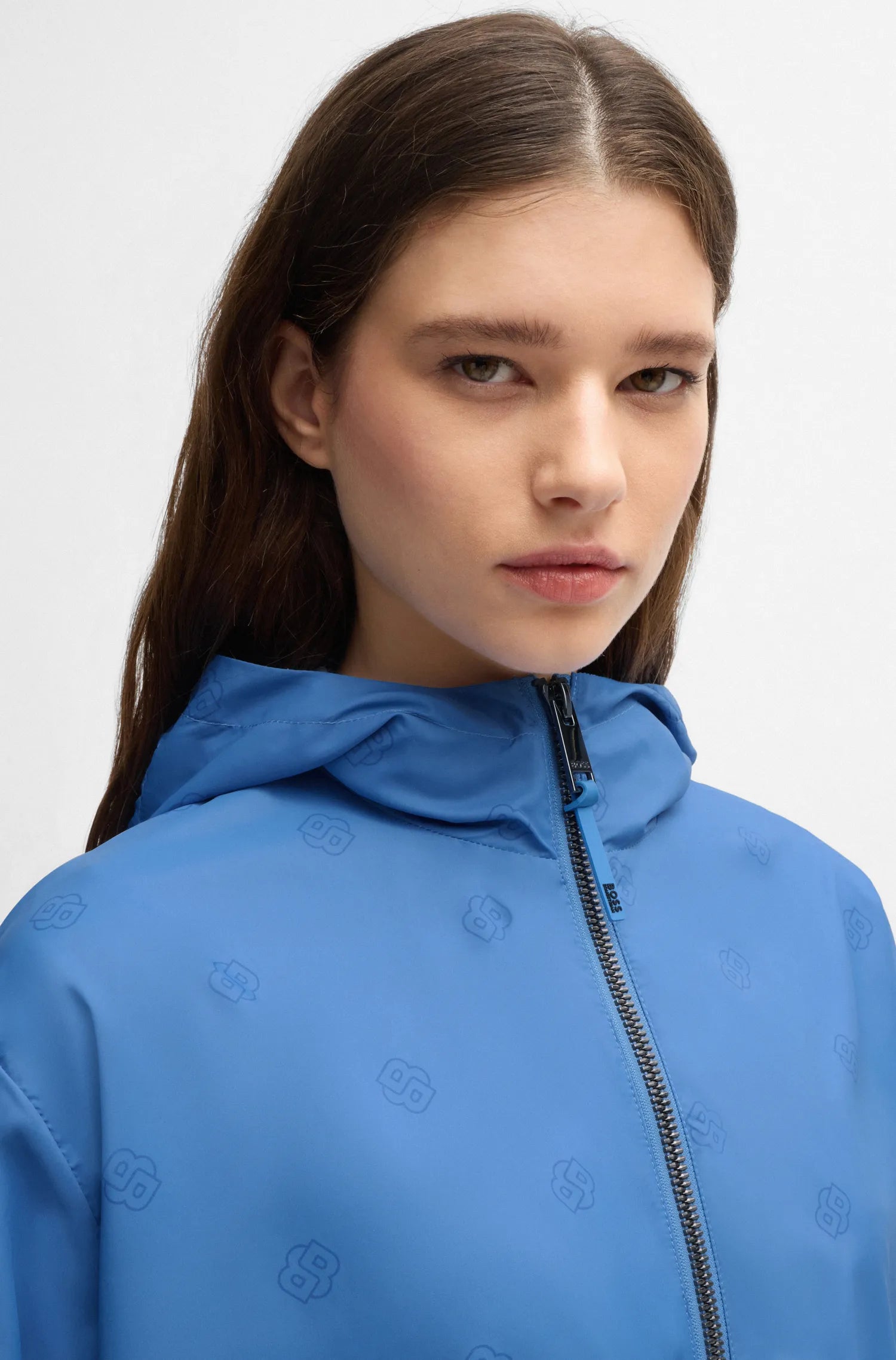 Riviera | A close-up shot of a model wearing a bright blue hooded jacket with a subtle tonal monogram pattern and a black zipper, set against a plain light gray background.