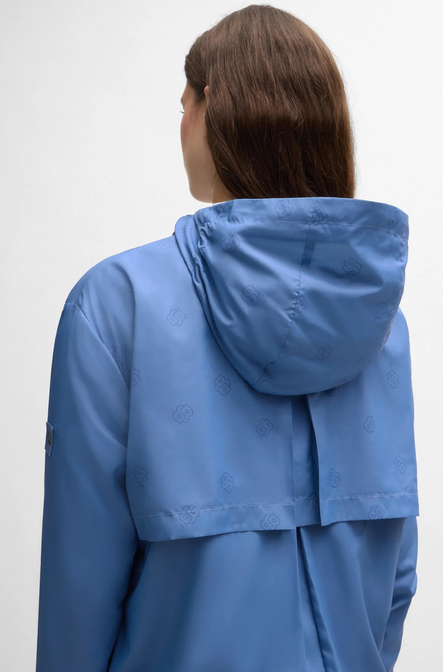 Riviera | A woman viewed from behind wearing a blue hooded jacket with a subtle tonal monogram pattern, set against a plain light gray studio background.