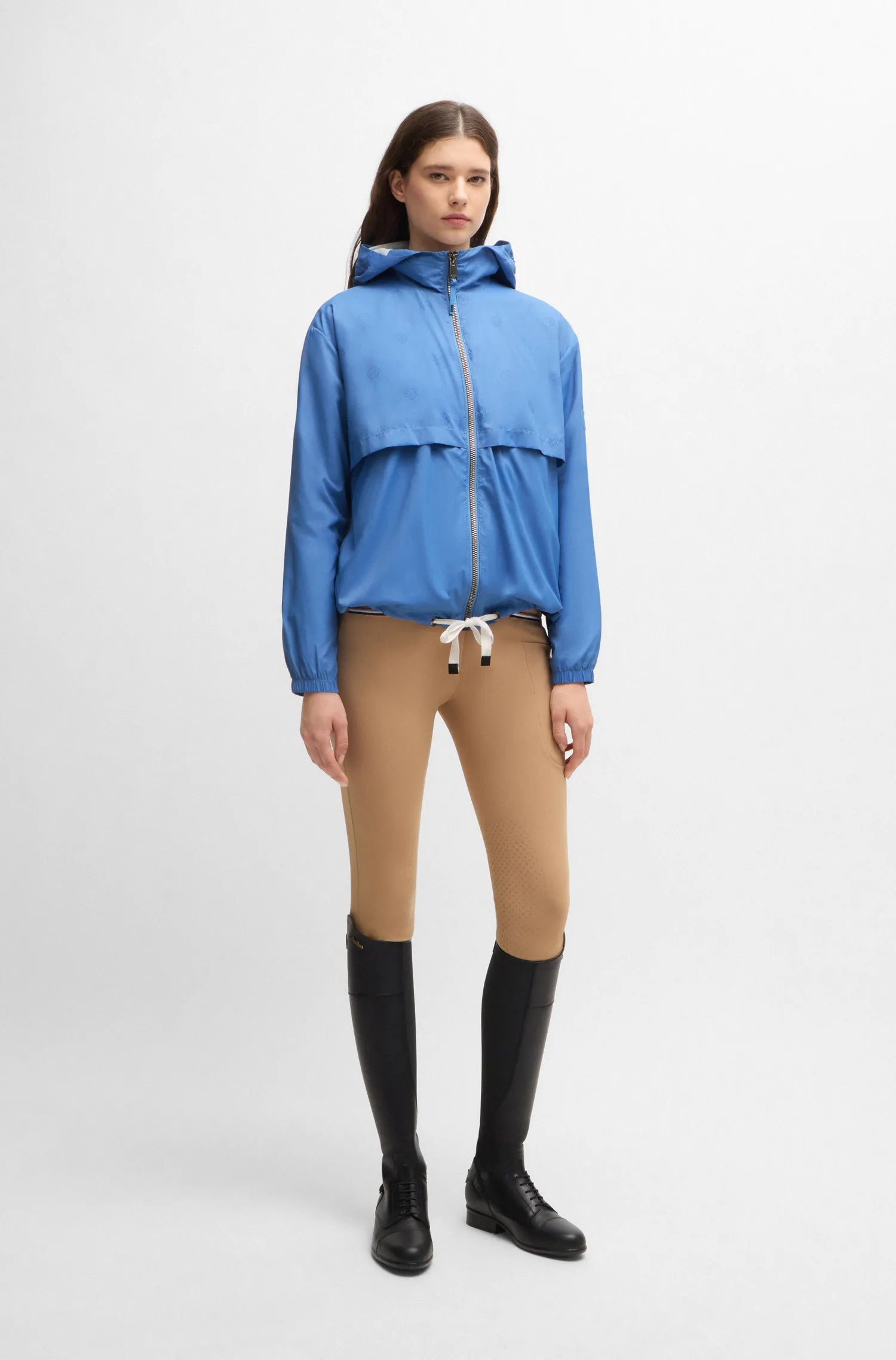 Riviera | A model stands against a light gray studio background wearing a bright blue hooded zip-front jacket, tan fitted pants, and black knee-high boots.