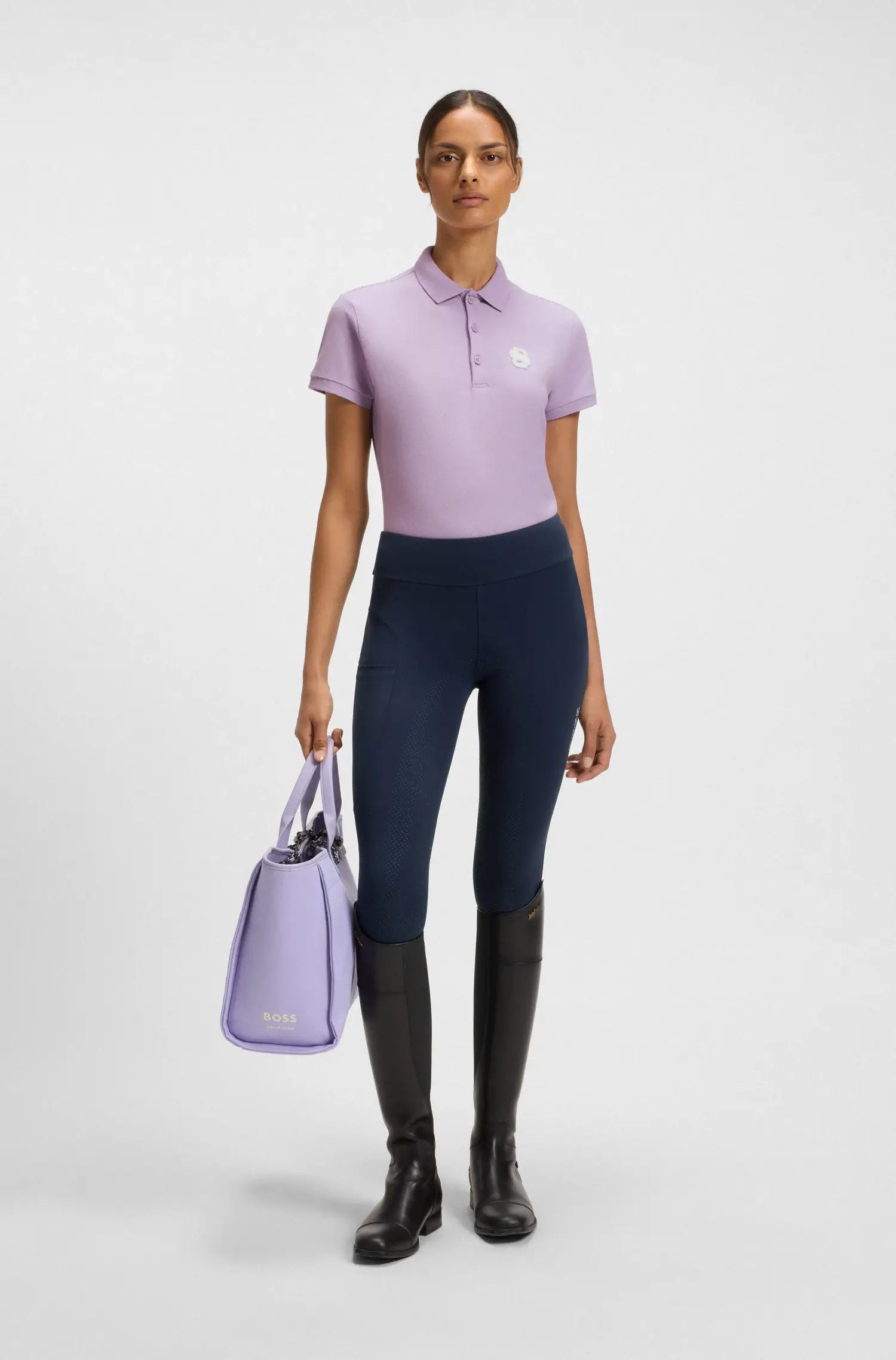 Orchid | Woman wearing a light purple polo shirt, dark blue leggings, and black riding boots, holding a matching purple bag on a white background.