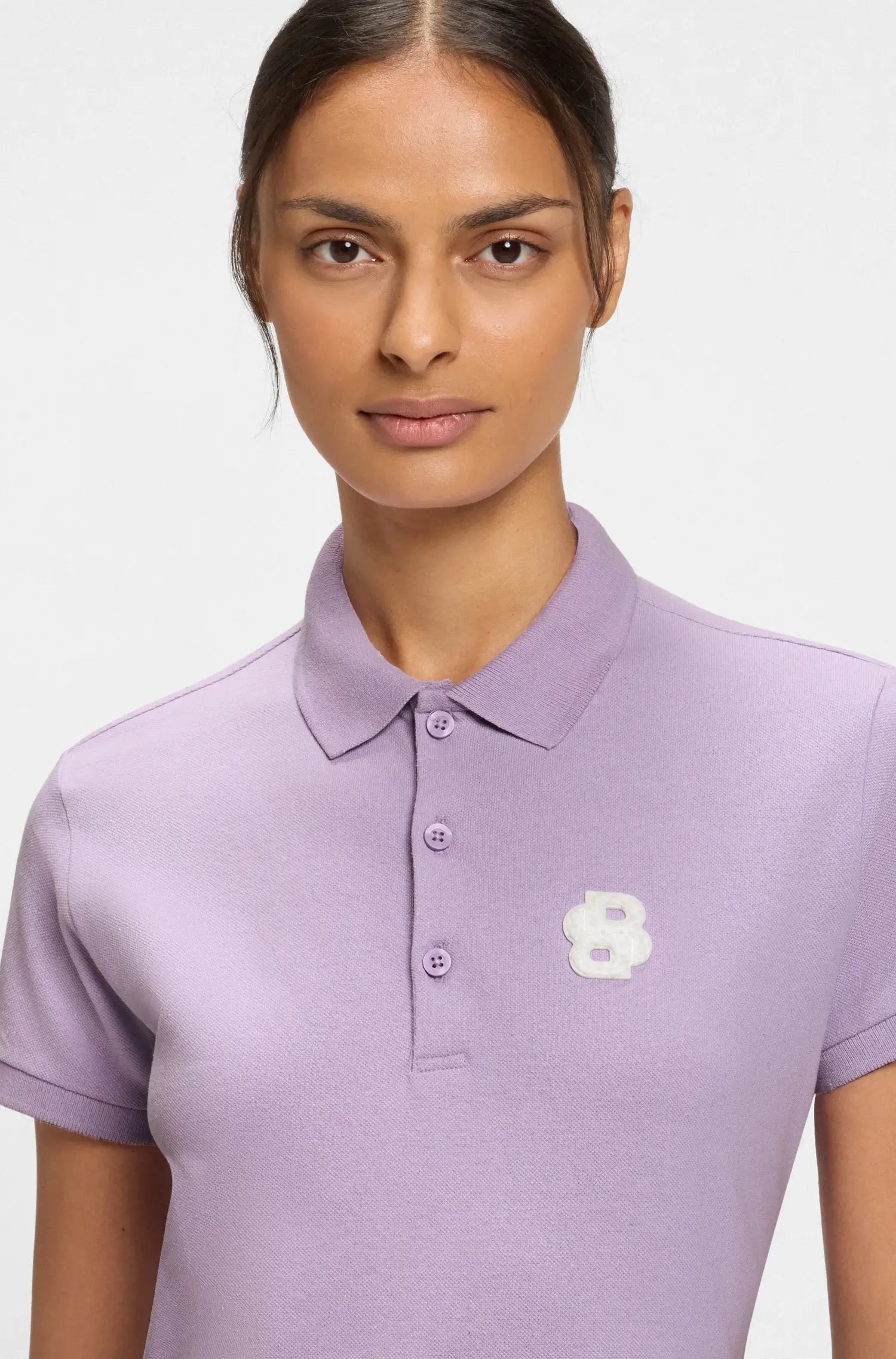 Orchid | Woman wearing a light purple polo shirt with a white flower logo on a plain background