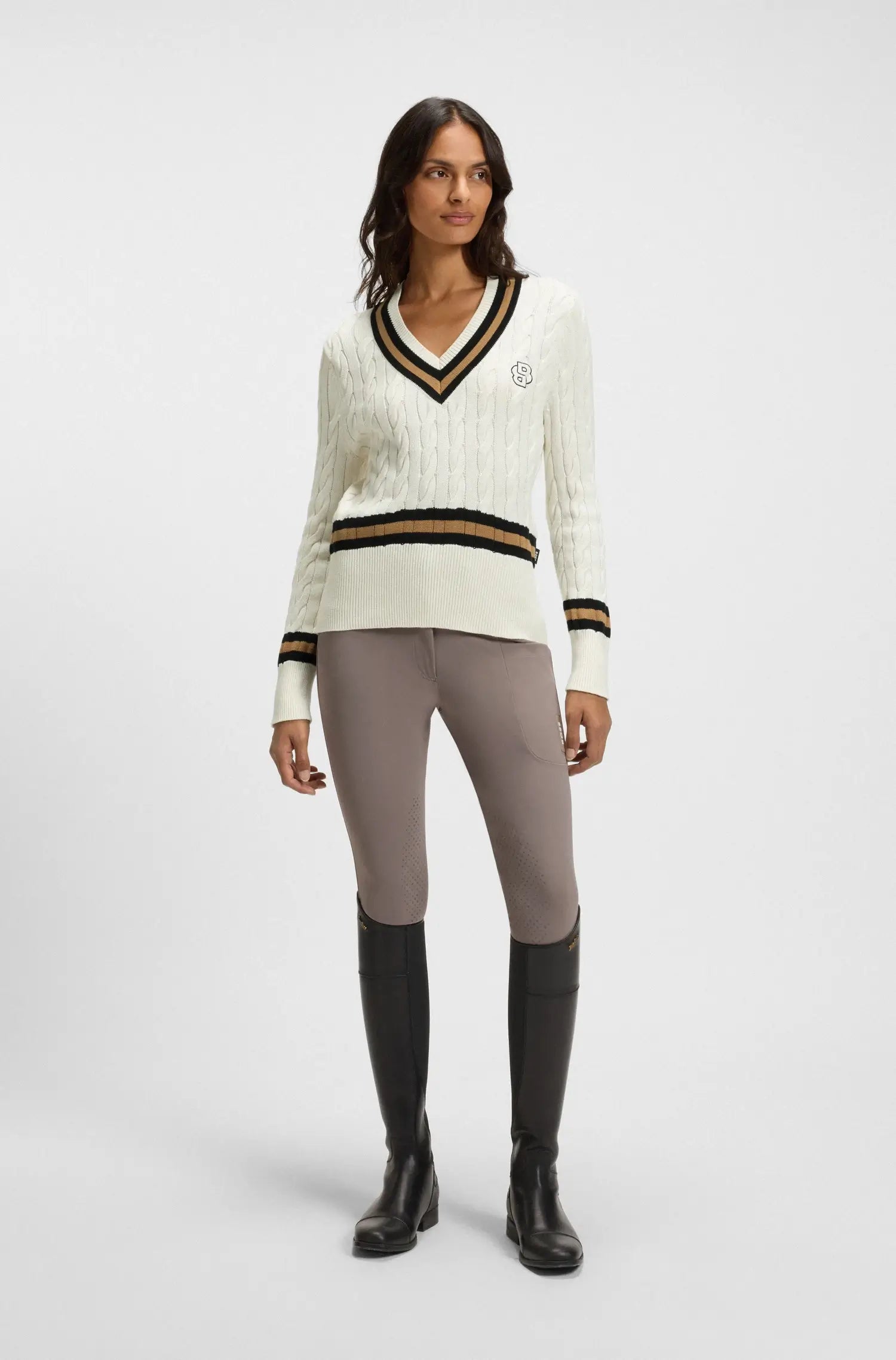 Soft Creme | Woman wearing a white sweater with black and gold accents, gray leggings, and black knee-high boots on a white background.
