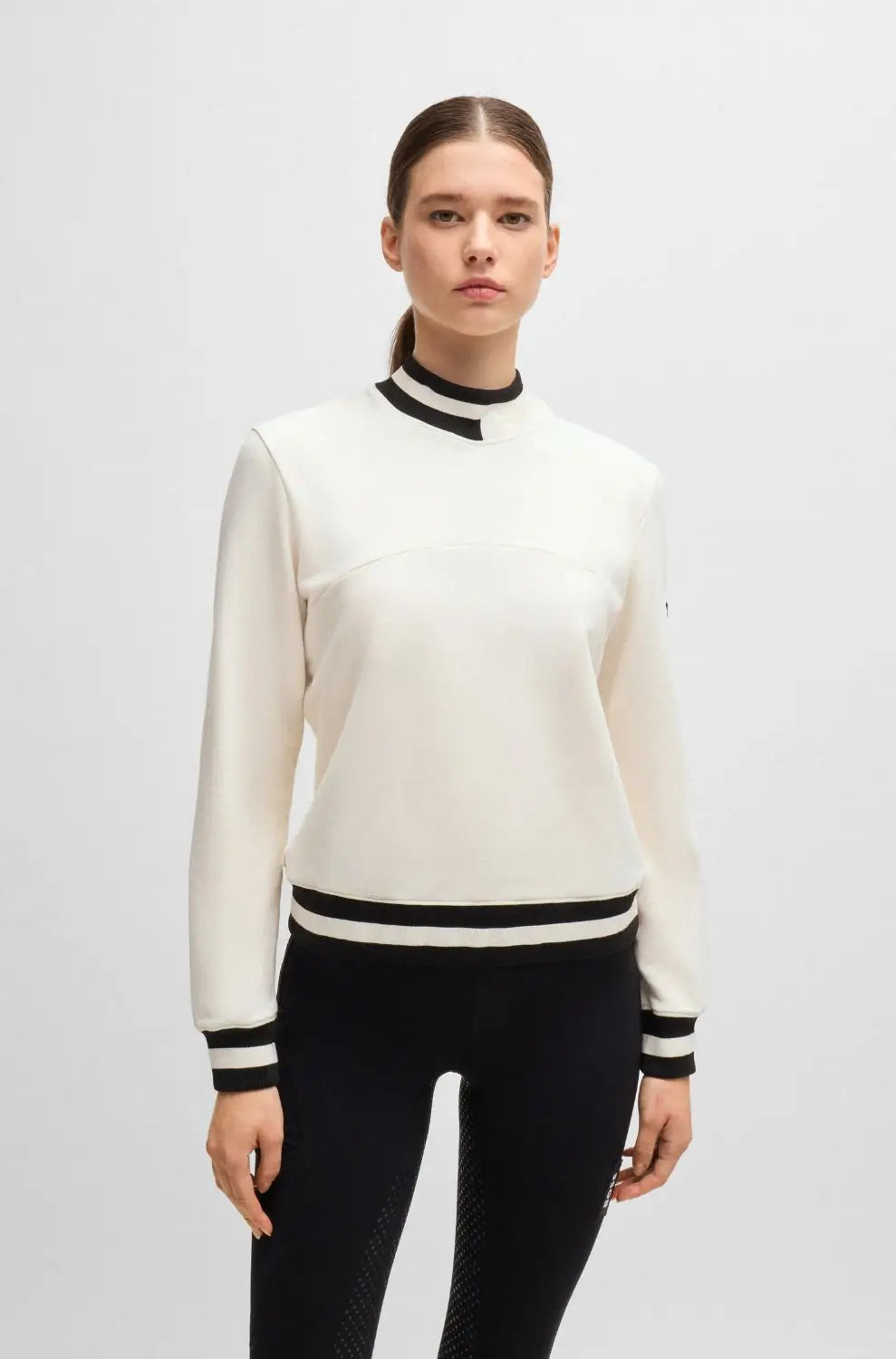 Pearl | A woman modeling a cream-colored mock neck sweater with black trim details at the collar, hem, and cuffs, paired with black pants, against a light gray studio background.