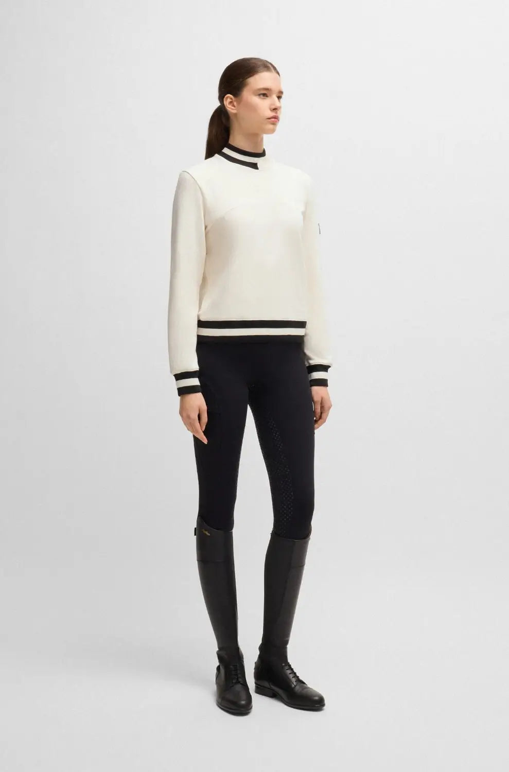 Pearl | A model stands against a light gray studio background wearing a cream mock-neck knit sweater with black and white stripes, black fitted pants, and black knee-high boots.