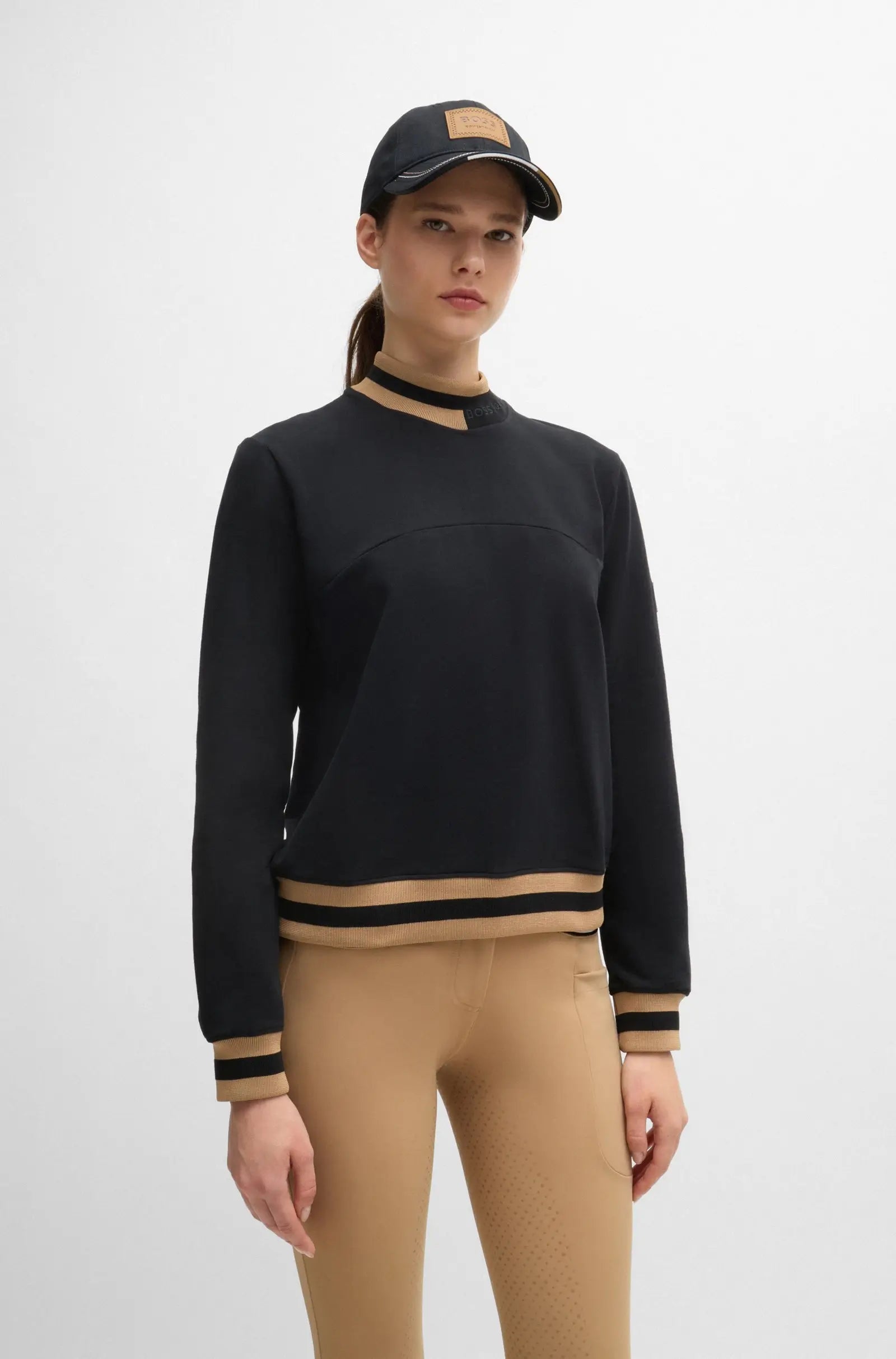Black | A model wearing a black mock-neck sweatshirt with beige and black striped ribbed trim, paired with beige leggings and a black baseball cap, against a plain light gray background.